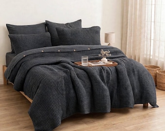 May include: A dark charcoal gray waffle-weave bedding set including a duvet cover with visible buttons and matching rectangular pillow shams. The textured fabric covers a bed frame, featuring a wooden tray with a glass of water on top.