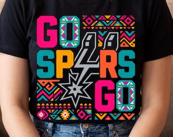 May include: Black t-shirt featuring the text "GO SPURS GO" in vibrant pink, orange, and teal. The design incorporates a silver lightning bolt and a star, with colourful geometric patterns. A casual wear item.