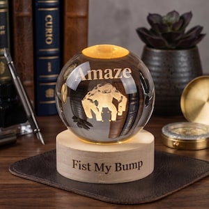 Puede incluir: Una esfera de cristal transparente sobre una base cilíndrica de madera clara. La esfera presenta la palabra Amaze grabada sobre un gráfico de un gorila. La base de madera tiene grabado el texto Fist My Bump en una fuente oscura.
