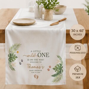 May include: A white table runner measuring 30 x 40 inches, featuring green tropical leaf illustrations, animal paw prints, and baby footprints. The text reads: A LITTLE wild ONE IS ON THE WAY! WELCOME TO Thomas's BABY SHOWER JUNE 10, 2026.