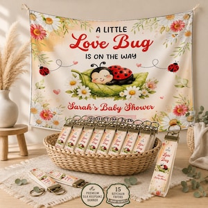 May include: A rectangular fabric banner featuring a ladybug on a leaf, daisy flowers, and the text A LITTLE Love Bug IS ON THE WAY and Sarah's Baby Shower. A woven basket contains multiple matching fabric keychains with the same design.