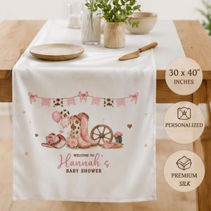 May include: A white table runner measuring 30 x 40 inches, featuring a printed design of pink cowboy boots, a hat, a wagon wheel, balloons, and floral accents. The text reads Welcome to Hannah's Baby Shower in pink script and block lettering.