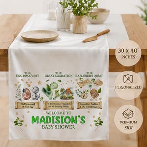 May include: A white table runner measuring 30 x 40 inches featuring dinosaur illustrations, gold stars, and text reading The Egg Discovery, The Great Migration, The Explorer's Quest, and Welcome to Madison's Baby Shower with personalized details.