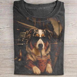 Könnte beinhalten: Dunkelgraues T-Shirt mit einem Hund, der eine Sonnenbrille trägt und ein Kaffeegetränk mit Strohhalm hält. Der Hund ist braun, schwarz und weiß, mit einer Stadtkulisse im Hintergrund. Das T-Shirt hat einen Vintage-Look.