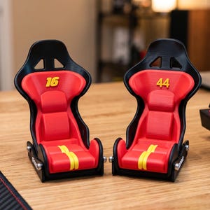 May include: Two miniature racing bucket seats featuring black frames and red cushions. The left seat displays the yellow number 16, while the right seat displays the yellow number 44. Both seats include yellow vertical stripes on the seat base.