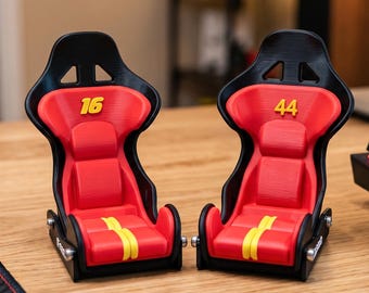 以下が含まれることがあります： 黒いフレームと赤いクッションを備えた2つのミニチュアレーシングバケットシート。左側のシートには黄色の数字16、右側のシートには黄色の数字44が表示されています。両方のシートの座面には黄色の縦縞模様が施されています。
