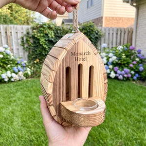 May include: A teardrop-shaped wooden butterfly house featuring a natural wood grain finish, three vertical entry slots, and a small circular glass dish attached to the front. The text "Monarch Hotel" is engraved on the upper front panel.