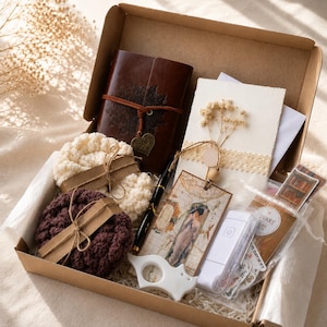 May include: A gift box containing a dark brown leather journal with a heart charm, two crocheted coasters in cream and dark brown, a black pen, a decorative card with dried flowers, a vintage-style bookmark, a white plastic tool, and stickers.