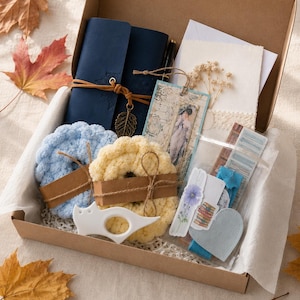 May include: A cardboard gift box containing a dark blue leather-bound journal with a leaf charm, two round crocheted coasters in light blue and yellow, a bookmark with a vintage illustration, a white cat-shaped tool, and various paper stationery items.