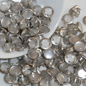 May include: A collection of silver-toned, round buttons with a pearlescent, slightly domed surface. The buttons are scattered, some spilling from a white, shallow dish. Each button has a metal shank on the back for sewing onto fabric.