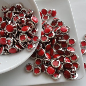 Puede incluir: Una colección de tachuelas rojas para tapicería con bordes de metal plateado y puntas. Las tachuelas están dispersas sobre una superficie blanca y en un cuenco blanco. Estos pequeños sujetadores redondos son adecuados para tapicería y manualidades.