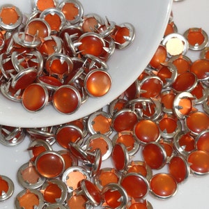 May include: A collection of orange-colored upholstery tacks with silver-colored metal rims. The tacks are scattered on a white surface, with some spilling from a white bowl. The orange tops have a glossy finish.