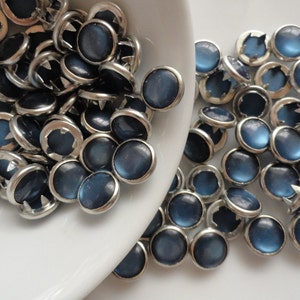 May include: A collection of round, dark blue buttons with silver-colored metal rims. The buttons have a slightly domed, matte finish and are scattered on a white surface, some in a white bowl.