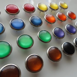 May include: Assortment of colorful round buttons with a silver metal rim. The buttons have a variety of colors including red, orange, yellow, green, blue, purple, and black.