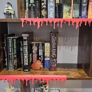 Pu&ograve; includere: Una libreria in legno con libri di Stephen King. I ripiani sono decorati con vernice rosa brillante gocciolante. I titoli includono "The Tommyknockers", "Doctor Sleep" e "Pet Sematary".