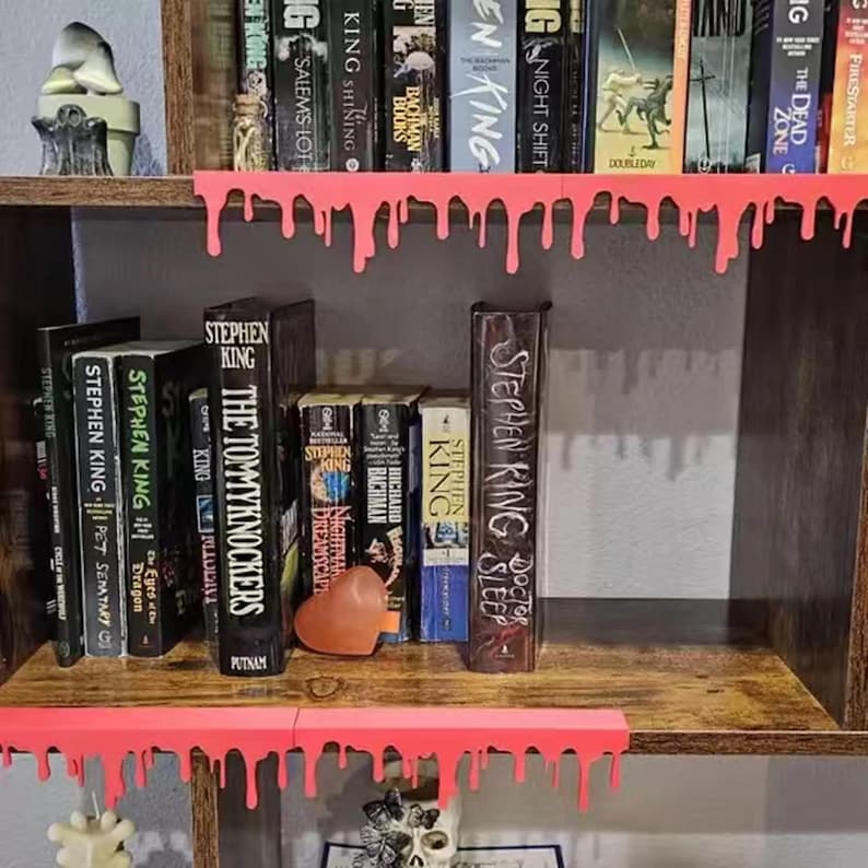 Pu&ograve; includere: Una libreria in legno con libri di Stephen King. I ripiani sono decorati con vernice rosa brillante gocciolante. I titoli includono "The Tommyknockers", "Doctor Sleep" e "Pet Sematary".
