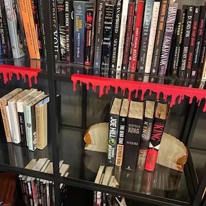 Pu&ograve; includere: Una libreria nera con una collezione di libri, alcuni con dettagli rossi. La libreria presenta un design unico con elementi rossi a forma di goccia. Sono visibili diversi libri di Stephen King.