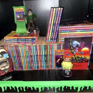 Pu&ograve; includere: Una collezione di libri e merchandising di Goosebumps. L'immagine presenta una variet&agrave; di libri con dorsi colorati, statuette e una mensola verde a forma di slime. I libri sono disposti su scaffali, creando un'esposizione della popolare serie horror per bambini.