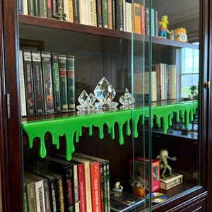 Pu&ograve; includere: Una vetrina in legno scuro con ante e ripiani in vetro. Il ripiano superiore presenta un design a goccia di melma verde brillante. Sculture di cristallo trasparente sono appoggiate sul ripiano. I libri sono visibili sui ripiani, insieme ad altri oggetti decorativi.