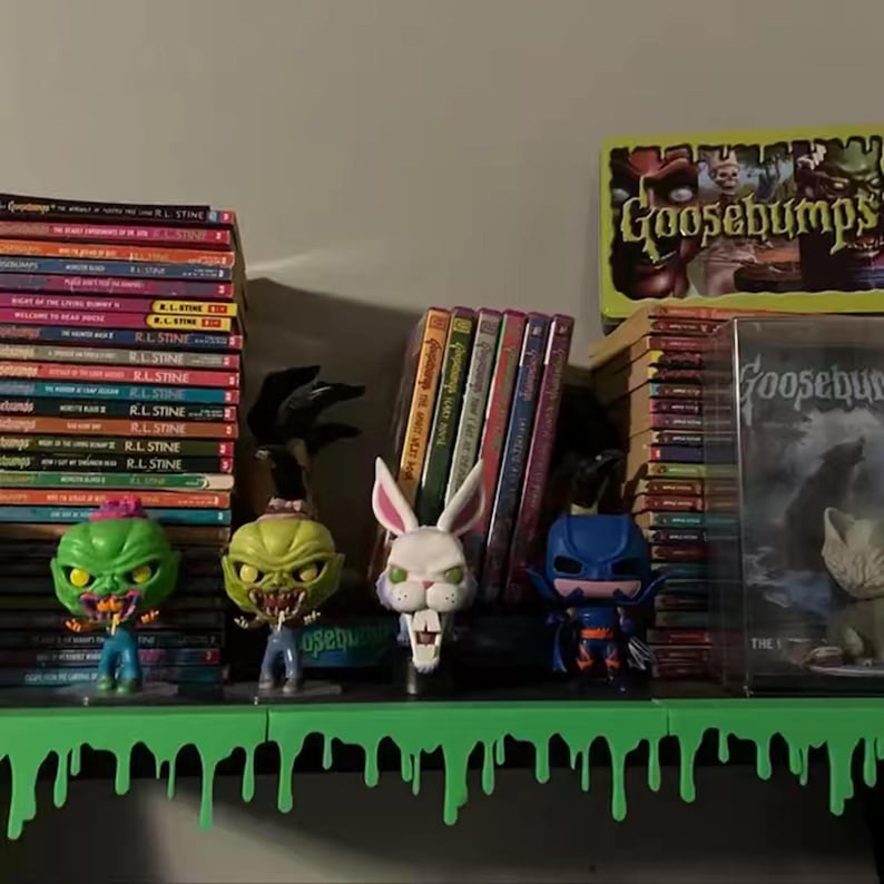 Pu&ograve; includere: Una mensola espone libri, DVD e statuette di Goosebumps. La mensola &egrave; dipinta con vernice verde gocciolante. Le statuette includono un mostro verde, un coniglio bianco e un supereroe blu. I libri sono impilati.