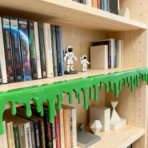 Pu&ograve; includere: Libreria in legno con vari libri, tra cui Goosebumps. Due statuette di astronauti sono su uno scaffale con un design verde brillante a goccia. Sono visibili anche forme geometriche e altri libri.