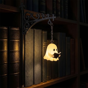 May include: A small, glowing, off-white ghost-shaped lamp holding a black book, suspended by a silver-toned chain from a black, ornate, scroll-work metal wall bracket. The lamp emits a warm, soft light against a background of dark books.