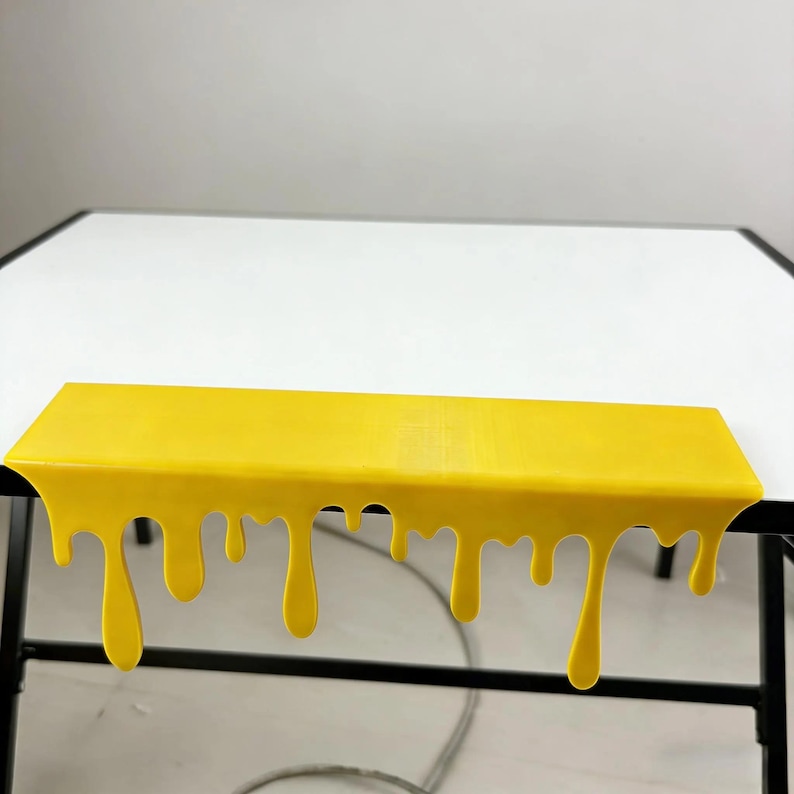 Pu&ograve; includere: Una mensola rettangolare giallo brillante con un design a goccia lungo il bordo inferiore. La mensola &egrave; posizionata su una superficie bianca, con gambe in metallo nero sottostanti. Il design ricorda la vernice che si scioglie o gocciola.