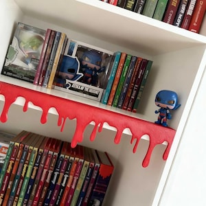 Pu&ograve; includere: Una libreria bianca con un bordo rosso gocciolante. La mensola espone vari libri e figurine da collezione, tra cui una figura di supereroe blu. Il testo "ATTACK OF THE MUTANT" &egrave; visibile su una scatola.