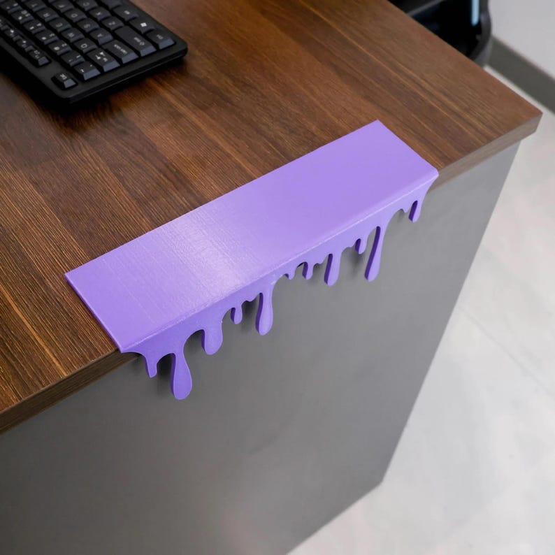 Pu&ograve; includere: Un proteggi-angoli per scrivania viola con un design a goccia di vernice. Il protettore rettangolare &egrave; montato sull'angolo di una scrivania in legno marrone. L'effetto goccia di vernice aggiunge un tocco artistico unico alla scrivania.