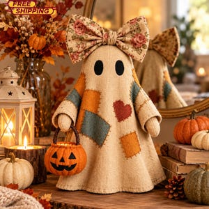 May include: A beige fabric ghost figurine featuring a large floral print bow on its head and patches in orange, blue, and brown. The ghost has two black oval eyes, a red heart patch, and holds a small orange jack-o'-lantern bucket in its hand.