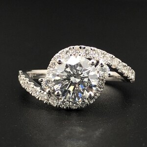 May include: A silver-toned ring featuring a large, round, faceted center stone surrounded by a halo of smaller, round, sparkling stones. The band is split and curves around the center setting, encrusted with additional small, round stones.
