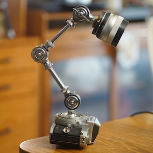 May include: A desk lamp constructed from a vintage silver and black film camera base with an adjustable articulated metal arm and a camera lens head. The lamp features multiple circular joints and a metallic finish, combining photographic equipment.