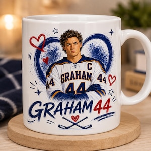 May include: A white ceramic mug featuring a graphic of a hockey player wearing a white jersey with the name GRAHAM and number 44. The design includes a blue heart, red hearts, stars, and the text GRAHAM 44 above two crossed hockey sticks.