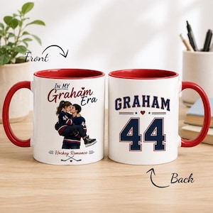 May include: Two white ceramic mugs with red interiors and handles. The front mug features an illustration of a couple in hockey jerseys with the text In My Graham Era and Hockey Romance. The back mug displays the name GRAHAM and the number 44.