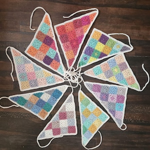 May include: A set of eight triangular crochet bunting flags arranged in a circular pattern. Each flag is composed of multiple small, multi-colored granny squares stitched together with white yarn borders and attached to white yarn ties.