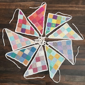 May include: A set of eight triangular crochet bunting flags arranged in a circular pattern. Each flag is composed of multi-colored square patches in various pastel and vibrant hues, featuring white crocheted borders and attached white yarn ties.