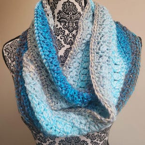 May include: A handmade, crocheted infinity scarf in shades of blue, gray, and white. The scarf features a textured stitch pattern and is displayed on a mannequin. The color gradient transitions from light to dark blue, creating a visually appealing accessory.