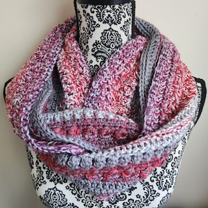 May include: A chunky crochet infinity scarf featuring a variegated yarn blend of vibrant red, soft pink, deep purple, and neutral gray tones. The accessory displays a textured stitch pattern with visible loops and rows, creating a thick, layered look.