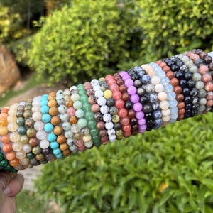 Könnte beinhalten: Eine Sammlung zahlreicher Perlenarmbänder, die in einer Reihe auf einem zylindrischen grauen Präsentationsständer angeordnet sind. Die Armbänder bestehen aus kugelförmigen Perlen in einer Vielzahl von Farben, darunter Grün, Orange, Gelb, Blau, Weiß, Rosa, Violett und Schwarz.