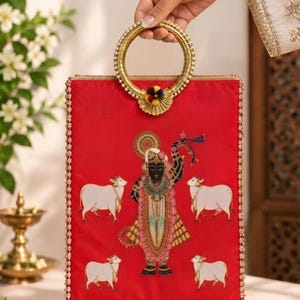May include: A rectangular red fabric handbag featuring a central illustration of a deity figure surrounded by four white cows. The bag has a circular gold-toned metal handle with a small multicolored pom-pom accent and gold beaded trim edges.