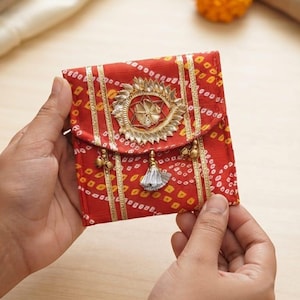 May include: A small square red fabric pouch featuring a white tie-dye pattern, gold vertical stripes, and a central circular gold embellishment. The flap is decorated with a small silver tassel and two gold-toned metal bells hanging from the edge.