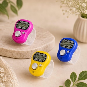 May include: Three digital tally counters with adjustable white silicone finger straps. Each device features a bright plastic casing in pink, yellow, or blue, a small LCD screen displaying the number 00025, a large silver count button, and a reset button.