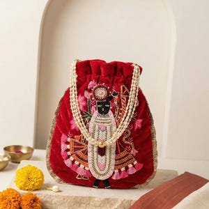 May include: A deep red velvet drawstring pouch featuring intricate beadwork and embroidery depicting a central figure. The bag is adorned with a double-strand pearl handle and gold-toned trim along the edges, showcasing detailed craftsmanship.