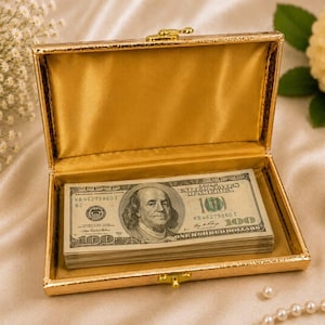 May include: A rectangular gold-colored box with a hinged lid and a metallic latch, featuring a smooth gold satin interior lining. Inside the box rests a stack of United States one hundred dollar bills, each clearly displaying Benjamin Franklin.