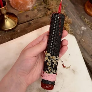 May include: A cylindrical black beeswax candle with a honeycomb texture, featuring a red wick at the top and a red wax base. The candle is decorated with small dried white flowers and a light pink string wrapped around the lower section.