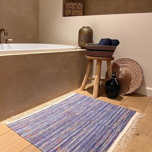Peut inclure: Tapis en tissu rayé bleu et blanc avec une bordure effilochée sur un sol en bois. Le tapis est dans une salle de bain, avec une baignoire, un tabouret en bois et des objets décoratifs.