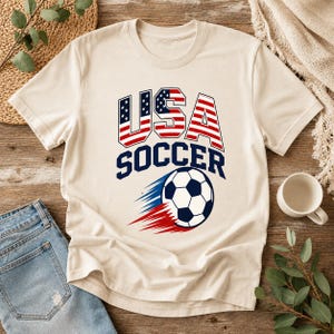May include: Cream-colored t-shirt featuring the text "USA SOCCER" in blue and red. The word "USA" is designed with the American flag. A soccer ball with blue and red speed lines is below the text. The shirt is displayed on a wooden surface.