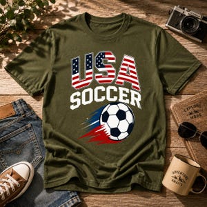 May include: Olive green t-shirt with "USA SOCCER" text and a soccer ball graphic. The text "USA" is designed with the American flag. The soccer ball has black and white panels with red and blue motion lines.