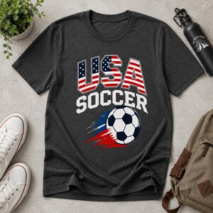 May include: Dark gray t-shirt with a USA Soccer graphic. The design features the word "USA" in red, white, and blue, with a soccer ball and the word "SOCCER" in white. The shirt is made of a soft material.
