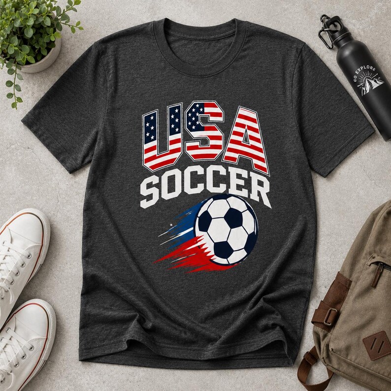 May include: Dark gray t-shirt with a USA Soccer graphic. The design features the word "USA" in red, white, and blue, with a soccer ball and the word "SOCCER" in white. The shirt is made of a soft material.